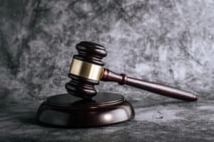 Gavel resting on sound block, symbolizing justice and legal authority. The gavel is made of dark wood with a gold band, set against a gray textured background.